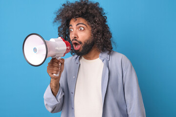 Young surprised curly Arabian man with megaphone in hands widens eyes making shocked grimace and...