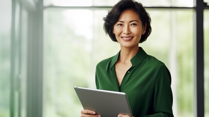 Smiling mid aged mature professional business woman in forest green blouse, 40s female executive or entrepreneur holding fintech tab digital tablet standing in office at work, large window copy space