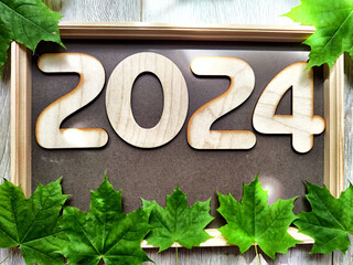 Wooden figures and numbers of the new year 2024 with frame and green maple leaves. Celebration with...