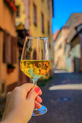 Woman s hand with a glass of white wine at an outdoor restaurant, blurred half timbered houses in Riquewihr, France, a village on Alsatian Wine Route