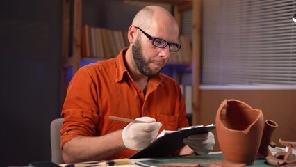 Bearded male archaeologist working late at night in office writing data on clipboard while sitting at desk working overtime