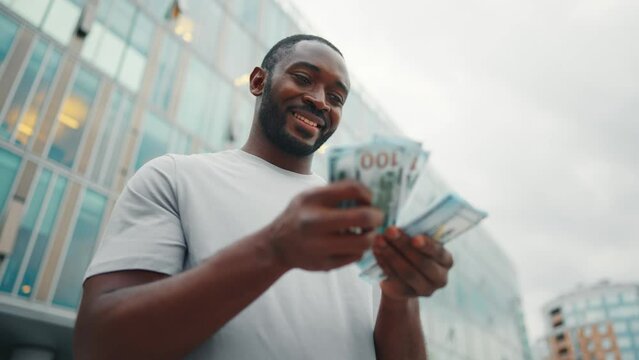 Man counts dollars banknotes on street. Contented smiling african american guy enjoying earned cash money in city business center, low angle view. Finances, wages concept. Entrepreneur, businessman.
