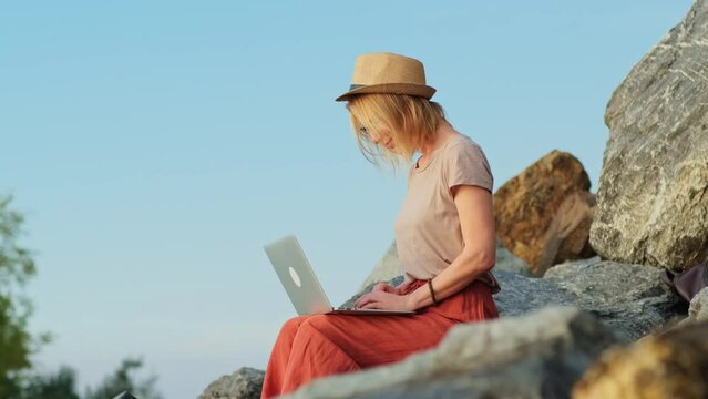 Side view of freelancer woman using a laptop while sitting on a high stone outdoor. Secret place for inspiration.