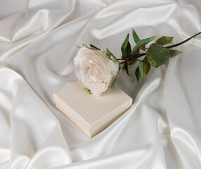 white rose lies on a gift box on a white satin draped background