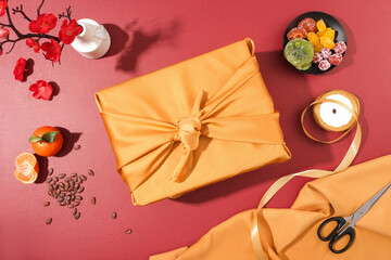 A gift box wrapped in yellow silk cloth, a plate of fruit jam, a roll of ribbon, melon seeds,...