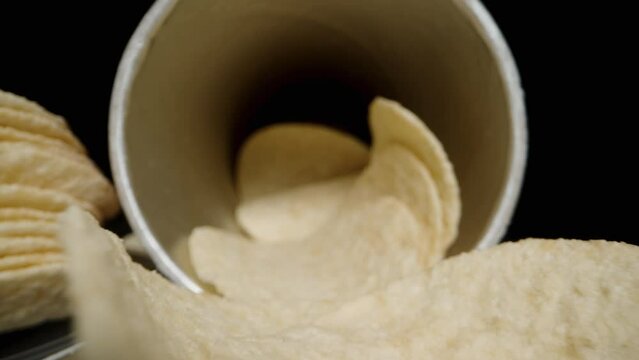Corn chips, hyperbolic paraboloid in shape, are scattered from a can onto the table against a black background. Dolly slider, close up.