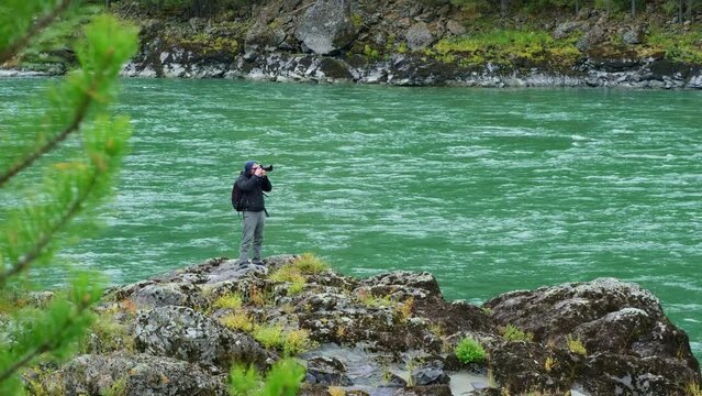 Male photographer taking photos of mountain river landscape while standing on the rocky shore. Traveler man and his camera on the rapid water stream background.