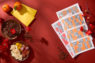 Top view of lotto set, a plate of melon seeds, salted dry apricot, tangerines, lucky money envelopes and peach blossoms on a red background. Tet game concept.