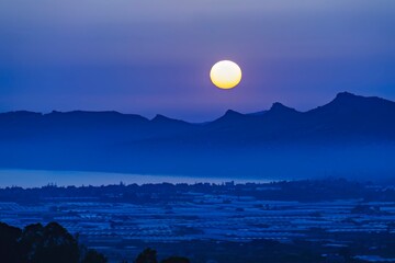 Full moon rising above serene blue mountains and silhouetted peaks. Full Moon Over Tranquil...