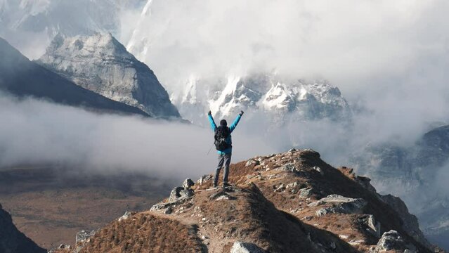 Tourist explorer with backpack hiking in mountains of Himalayas. At top of mountain he raises arms up celebrating achievements and success of ascent. Trekking through Nepal to Everest Base Camp