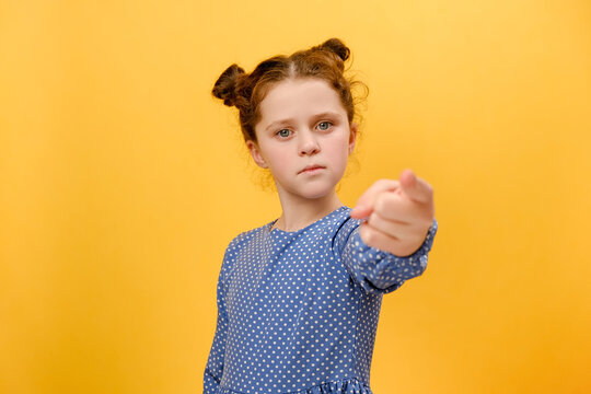 Portrait of serious little caucasian girl child pointing at camera, looking angry, making choice, choosing guilty, posing isolated over plain yellow color background wall in studio with copy space