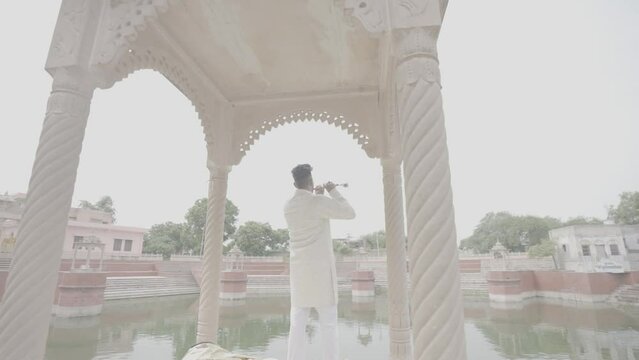 An Indian man's soulful flute resonates along the tranquil banks of the pond, weaving a melody that echoes the essence of nature and tradition.4k footage