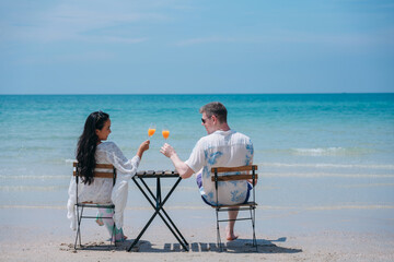 Obraz premium Happy and loving young couple drink orange juice at tropical beach. Couple Enjoying Fun and Romance by the Ocean.