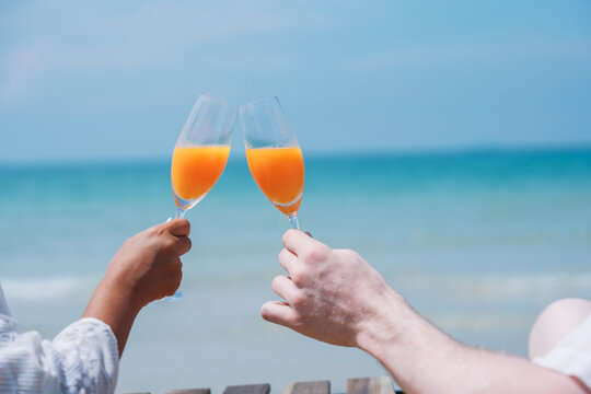 Happy And Loving Young Couple Drink Orange Juice At Tropical Beach. Couple Enjoying Fun And Romance By The Ocean.