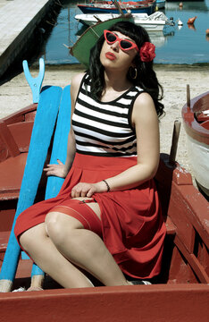 Harbor pin-up: Red skirt, stripes, retro glasses. Red heels, secrets beneath: garter belt, nude sheer stockings. Reimagined vintage elegance. 