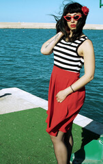 Port harbor pin-up: Red skirt, stripes, retro glasses. Red heels, intriguing secrets beneath: garter belt and sheer stockings. Revisited vintage elegance.