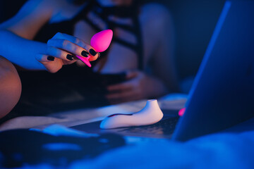 Beautiful woman lies in bed holding clitoral vibrator and watching porn on laptop, neon color. Women has Internet sex chat online - obrazy, fototapety, plakaty