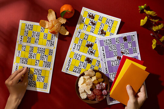 The lotto sheets are displayed on a red background with tangerines and salted dry apricot plates. A hand is holding lucky money envelopes. Top view.