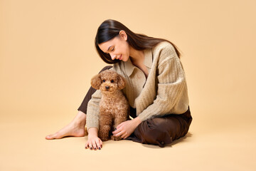 Young attractive lady sitting on floor with her lovely playful purebred dog, poodle against beige color studio background. Concept of animal, pet lover, friendship, domestic life.