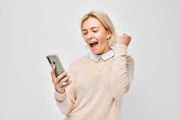 Fototapeta premium Portrait of young blond woman with mobile phone in hand celebrates victory, winning money in casino. Person with smartphone isolated on white background.