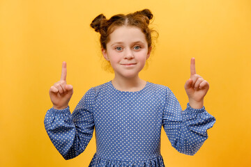 Portrait of cute funny little kid girl pointing index fingers up, posing isolated over yellow color background wall in studio. Childhood lifestyle concept. Mock up copy space for promotion content