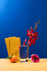 A fresh orchid is placed in a glass, two yellow envelopes, a gerbera flower and a tangerine are...