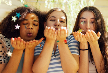 Close up of three cute happy teenage girls of different nationalities blowing star shaped confetti from their hands. - obrazy, fototapety, plakaty