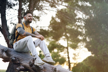 Smiling tattooed model man resting sitting on tree trunk in forest at sunset with back light