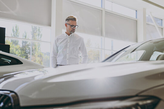 Adult rich serious man customer male buyer client he wears classic shirt look on hood choose auto want to buy new automobile in car showroom vehicle salon dealership store motor show. Sales concept.