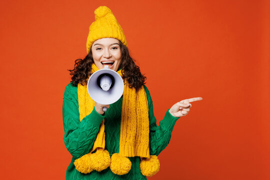 Young happy woman she wear green knitted sweater yellow hat scarf hold in hand megaphone scream announces discounts sale Hurry up point aside isolated on plain orange red background Lifestyle concept
