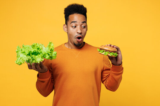 Young shocked man wear orange sweatshirt casual clothes hold lettuce salad burger choose what to eat isolated on plain yellow background. Proper nutrition healthy fast food unhealthy choice concept.