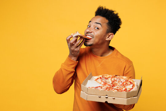 Young happy man wear orange sweatshirt casual clothes hold eating italian pizza in cardboard flatbox isolated on plain yellow background. Proper nutrition healthy fast food unhealthy choice concept.