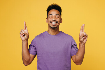 Young man of African American ethnicity he wears purple t-shirt casual clothes wait special moment, keep fingers crossed, make wish, eyes closed isolated on plain yellow background. Lifestyle concept.