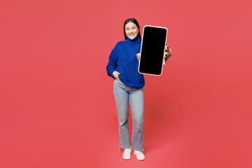 Full body young woman of Asian ethnicity she wear blue sweater casual clothes hold in hand use show mobile cell phone with blank screen workspace area isolated on plain pastel pink background studio.