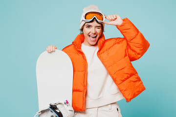 Young woman wears warm padded windbreaker jacket hat ski take off goggles mask hold snowboard look camera wink travel rest spend weekend winter season in mountains isolated on plain blue background. © ViDi Studio