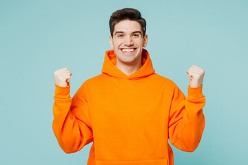 Young happy man he wears orange hoody casual clothes doing winner gesture celebrate clenching fists say yes isolated on plain pastel light blue cyan color background studio portrait Lifestyle concept