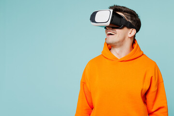 Young smiling happy man he wears orange hoody casual clothes watching in vr headset pc gadget look aside isolated on plain pastel light blue cyan color background studio portrait. Lifestyle concept.