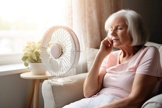 Exhauster older retired woman sitting at electric fan with propeller, enjoying cool fresh air flow, feeling relief, sitting on home sofa.