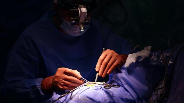 Professional neurosurgeon working in the dark surgery room with highlighted operational spot. Doctor uses metal forceps and thread to sew the patient.