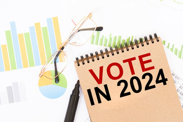 In a craft colour notebook is a VOTE IN 2024 inscription, next to pencils, glasses, graphs and...
