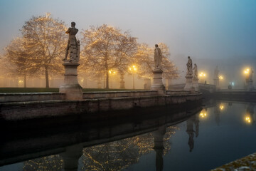 Padua, Italy - December 14, 2023: Prato della Valle, statues and trees with Christmas lights immersed in fog. - 699595047