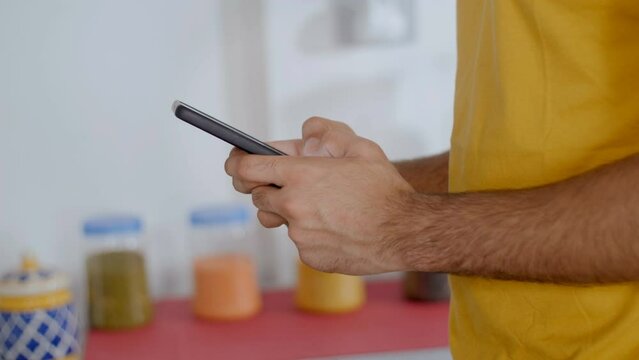 Close up of an Indian male using smartphone and typing . A young male in a yellow T-shirt sending text messages from mobile  standing near his kitchen area
