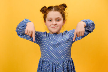 Portrait of pretty smiling little girl wearing blue dress pointing down place for commercial idea, looking at camera, posing isolated over yellow color background wall in studio. Look, advertise below