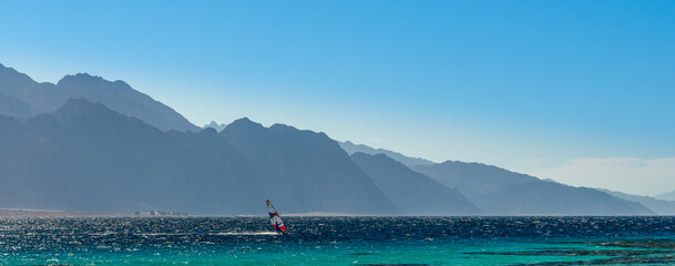 surfer rides in the Red Sea against the backdrop of high rocky mountains and a blue sky with clouds...