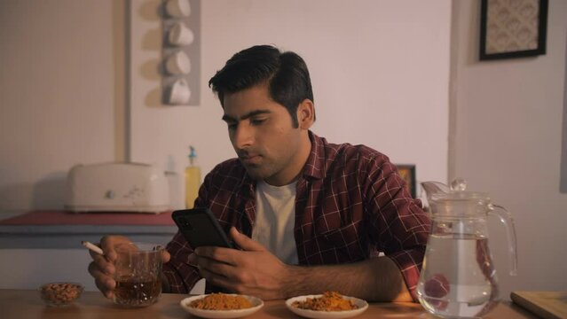 Drunk Indian man checking his mobile while sitting alone at home - alcohol addiction  drug addiction. Young unhappy male drinking and smoking while relaxing after working day - alcoholism problem  ...