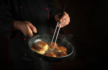 A cook in a black uniform fries delicious pancakes for breakfast. Hot frying pan in the chef hand and a fork. Dark place for menu or recipe.