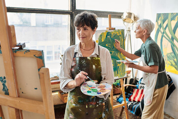brunette mature woman looking at easel while female friend painting in art workshop, creative hobby