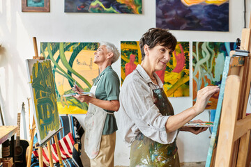 Fototapeta na wymiar joyful stylish mature women in aprons pointing on easels in art workshop, friendship and creativity