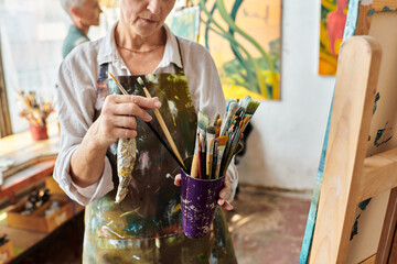 cropped view of mature woman choosing paintbrushes near female friend painting in art workshop