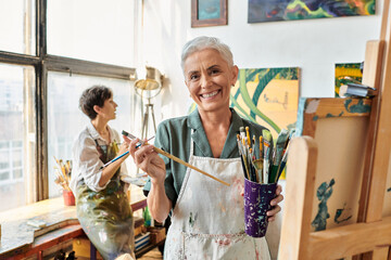 joyful mature woman with set of paintbrushes near female friend in art workshop, creative hobby